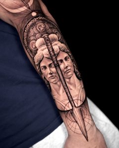 Detailliertes Tattoo mit zwei klassisch-skulpturalen Gesichtern, durchzogen von einer Speerspitze und umgeben von geometrischen Mustern