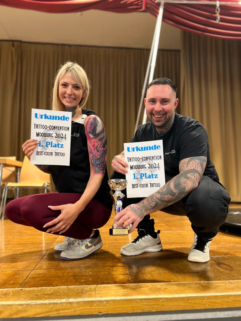 Der Besitzer von Skyline Tattoo posiert mit dem Mädchen, das er tätowiert hat. Beide halten die Urkunde in der Hand, und er hält den Pokal für den ersten Platz. Nummer eins an diesem Wochenende in Moosburg, Bayern.