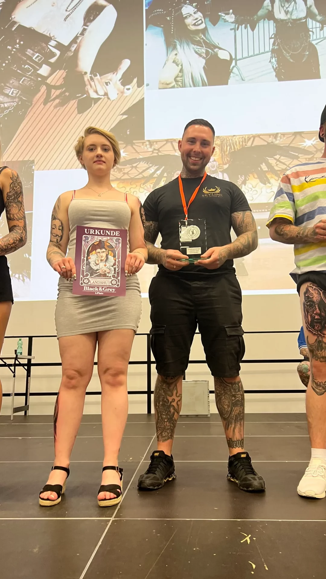 Der Besitzer von Skyline Tattoo posiert mit dem Mädchen, das er tätowiert hat. Sie hält die Urkunde in der Hand, er den Preis für den ersten Platz.