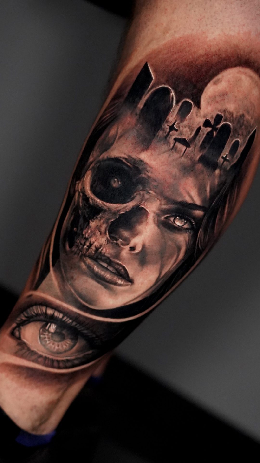 Realistisches Unterschenkel Tattoo mit halb weiblichem Gesicht und halb Totenkopf, kombiniert mit Friedhof, Grabsteinen und Vollmond im Hintergrund, darunter ein detailliertes Auge als zusätzliches Element, gestochen im Black & Grey Realismus Stil mit weichen Übergängen und starken Kontrasten, symbolisches Motiv für Leben und Tod, Vergänglichkeit und innere Dualität - gestochen bei Skyline Tattoo Bayern