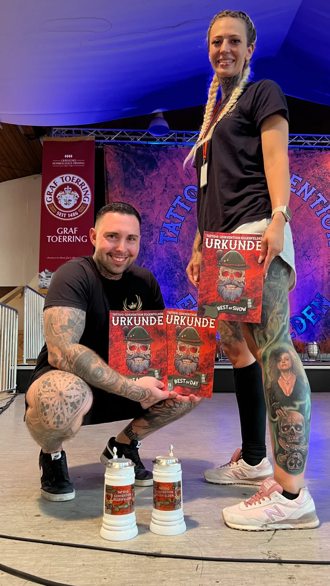 Der Besitzer von Skyline Tattoo posiert mit dem Mädchen, das er tätowiert hat. In der Hand halten sie Urkunden.