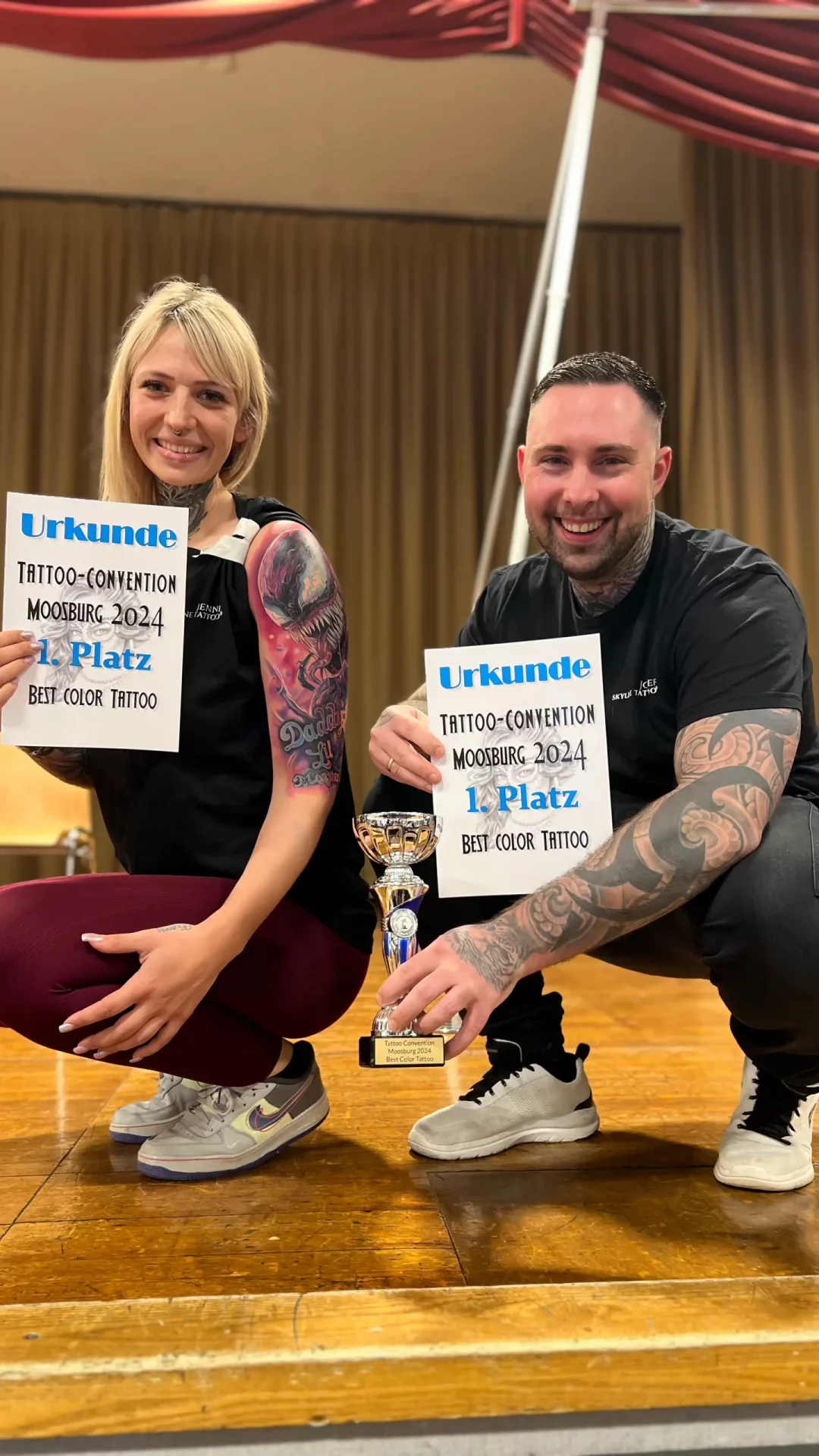 Der Besitzer von Skyline Tattoo posiert mit dem Mädchen, das er tätowiert hat. Beide halten die Urkunde in der Hand, und er hält den Pokal für den ersten Platz.