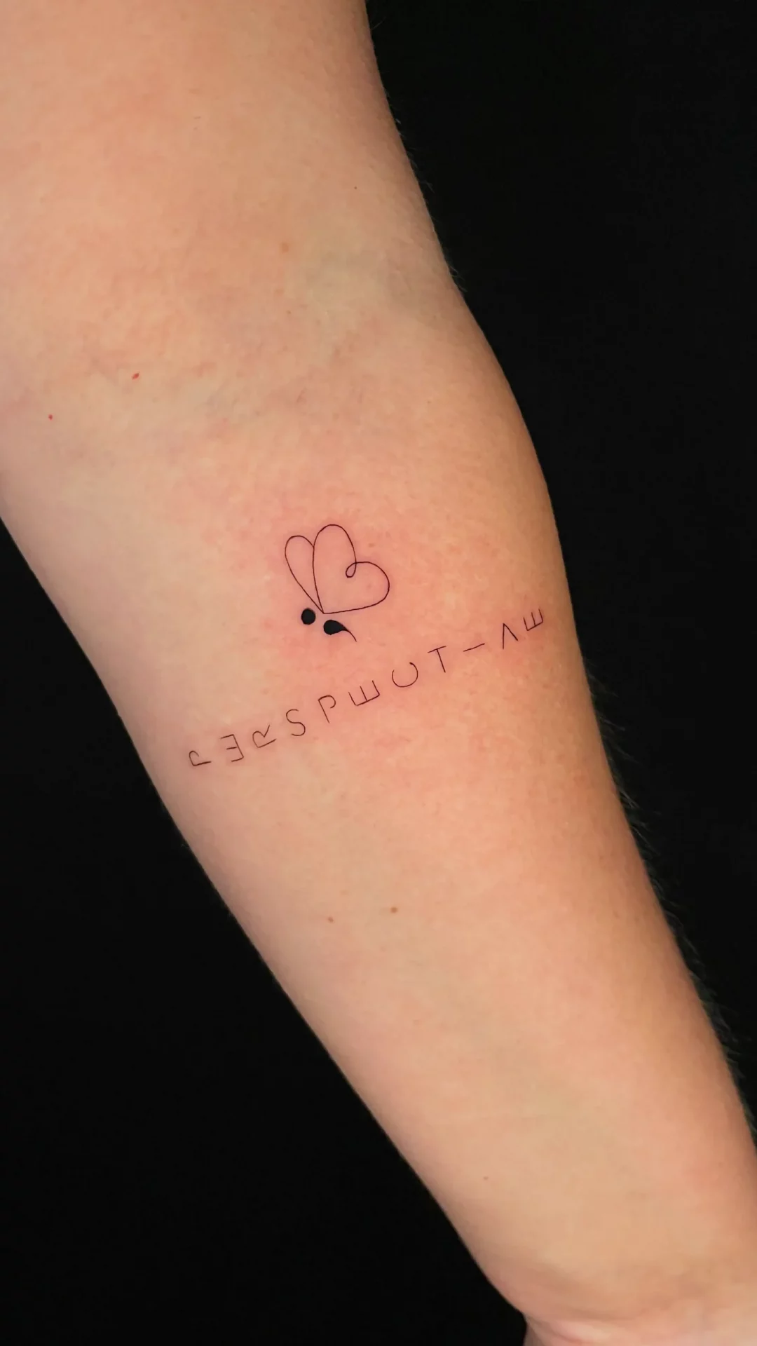 Fineline-Tattoo eines Schmetterlings, dargestellt mit einem Semikolon und einem Herz anstelle der Flügel, darunter der Schriftzug „perspective” mit verdrehten und gedrehten Buchstaben.